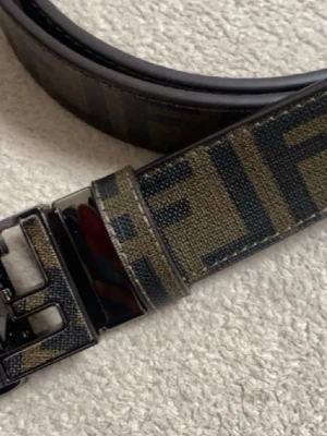 Svart och brun Fendi bälte - Snyggt bälte från Fendi i svart och brunt med klassiskt Fendi-mönster. Bältet har en stilren design med diskreta ränder och är tillverkat i skinn. Perfekt accessoar för att lyfta din outfit.