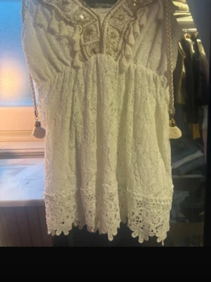Vit spetsklänning med broderi - Söt vit top i spets med broderade detaljer och paljetter vid halsringningen. Toppen har band  med tofsar och vackra volangdetaljer nertill. Perfekt för sommarens alla tillfällen.