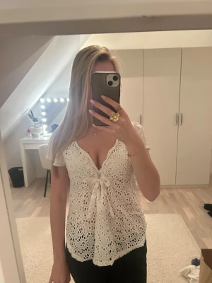 Vit spetsblus med knyt framtill - Söt vit blus i spets med v-ringning och knyt framtill. Blusen har korta ärmar och fina broderade detaljer som ger en romantisk känsla. Perfekt för dig som gillar feminina toppar med unik design.