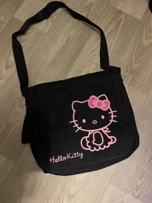 Hello Kitty svart axelväska - Säljer en svart axelväska med justerbar rem och stort Hello Kitty-tryck i rosa på framsidan. Perfekt storlek för en laptop.