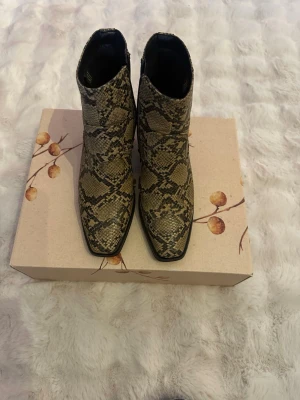 Ormmönstrade Cowboy boots med klack - Säljer ett par snygga cowboy boots från XTI i beige och svart ormmönstrat konstläder. Boots med spetsig tå, låg klack och dragkedja på sidan. Perfekta för dig som vill sticka ut med en trendig westernstil. Använda ett fåtal gånger 