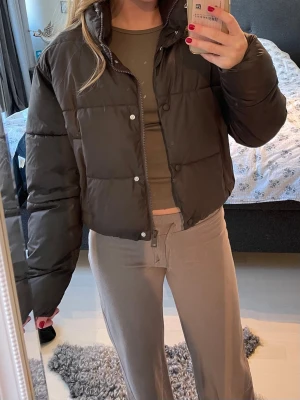 Brun pufferjacka med hög krage - Säljer en brun pufferjacka med hög krage och tryckknappar framtill. Jackan har en boxig, croppad passform och breda kanaler som ger en riktigt puffig look. Perfekt för kalla dagar när du vill vara både varm och trendig.