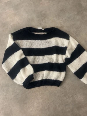 Randig stickad tröja i svart och vitt - Säljer en fluffig stickad tröja med breda svarta och vita ränder. Tröjan har rund halsringning, ballongärmar och ribbade muddar vid ärmslut och nederkant. Perfekt för dig som gillar en avslappnad och trendig look.