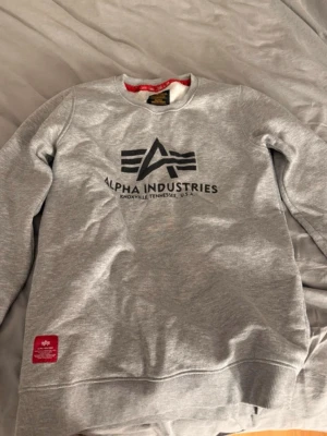 Grå sweatshirt från Alpha Industries - Snygg grå sweatshirt från Alpha Industries med stor svart logga och text på bröstet. Tröjan har rund halsringning, ribbade muddar vid ärmslut och nederkant samt en röd detalj på nederkanten. Perfekt för en avslappnad streetwear-look.