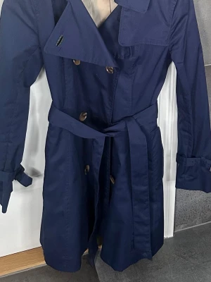 Mörkblå trenchcoat med bälte - Stilren mörkblå trenchcoat med dubbelknäppning, klassisk krage och knytskärp i midjan. Jackan har långa ärmar med spännen vid ärmsluten och stora knappar framtill. Materialet ser ut att vara bomull eller bomullsblandning. Perfekt för en trendig och tidlös look.