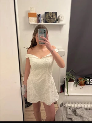 Vit offshoulder kortklänning - Söt vit kortklänning med offshoulder-modell och lätt utsvängd kjol. Klänningen har ett diskret mönster i tyget och en figurnära passform upptill. Perfekt för sommarens alla tillfällen och student. 