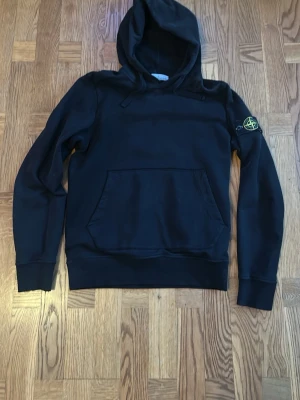 Svart hoodie från Stone Island - Säljer en svart hoodie från Stone Island med klassisk patch på vänster ärm och stor känguruficka framtill. Hoodien har dragsko i huvan och ribbade muddar vid ärmslut och nederkant. Tillverkad i mjuk bomull för skön komfort.