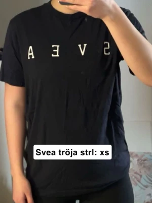 Svart t-shirt från Svea XS - Svart t-shirt från Svea i storlek XS med vit text på bröstet. Klassisk rund halsringning och korta ärmar. Tillverkad i mjuk bomull. Fint skick förutom skrynklig då den inte används men går att fixa me en tvätt eller strykning