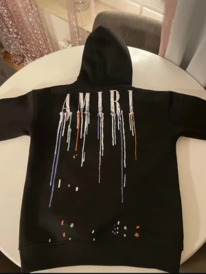 Svart hoodie från Amiri med tryck - Svart hoodie från Amiri med stor huva och känguruficka. På ryggen och bröstet finns AMIRI-logga med färgstänk och drip-effekt i vitt, blått och orange. Några färgdetaljer finns även nedtill. Snygg streetwear vibe och riktigt unik design.