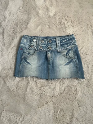 Blå jeanskjol med knappar - Säljer en blå kort jeanskjol med slitna detaljer och rå kant. Kjolen har sex stora silverfärgade knappar framtill och två fickor. Perfekt för dig som gillar en avslappnad och trendig stil. Pris kan diskuteras 