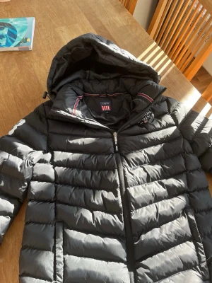 Svart dunjacka från U.S. Polo Assn. - Snygg svart dunjacka från U.S. Polo Assn. med huva och dragkedja framtill. Jackan har quiltad design, två sidofickor och logga på bröstet. Perfekt för kalla vinterdagar och riktigt skön att bära. Går att prata pris