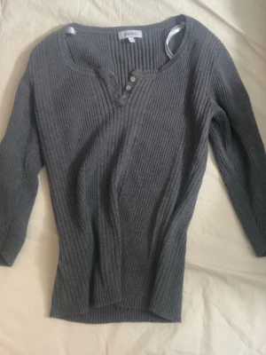 Mörkgrå ribbstickad topp  - Mörkgrå ribbstickad topp från Papaya med trekvartsärm och knappar vid halsringningen. Använd 1 eller 2 gånger. Tröjan har en smickrande, kroppsnära passform och bred rund hals. Perfekt för lager på lager och enkel att styla till olika looks.