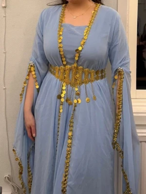 Ljusblå kurdisk klänning  - Ljusblå kurdisk klänning med guldiga paljettdetaljer och mynt längs kanterna. Klänningen har vida ärmar och ett elegant, flytande fall. 