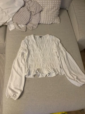 Vit smockad blus med ballongärmar - Trendig vit blus med smockad front och rygg, samt v-ringning. Blusen har långa ballongärmar med resår vid handlederna och en lätt croppad passform. Tillverkad i ett tunt, mjukt tyg som ger en luftig känsla. Perfekt för en stilren och modern look. Storlek S