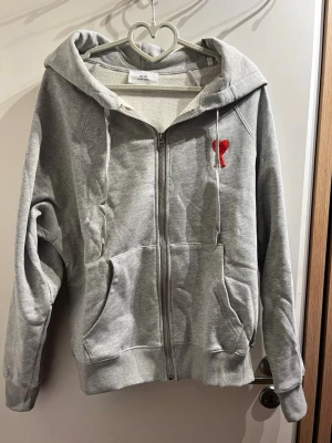 Grå zip hoodie från Ami Paris - Snygg grå hoodie från Ami Paris med dragkedja, stor huva och fickor framtill. Tillverkad i mjuk bomullsmix och har Ami-loggan i rött broderad på bröstet. Perfekt för en chill och avslappnad stil.