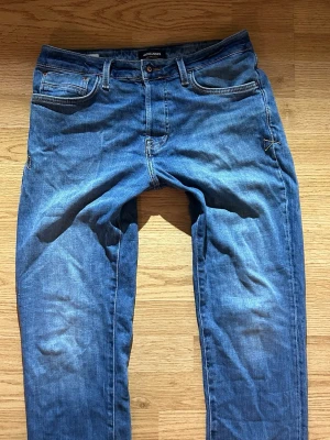 Blå jeans från Jack & Jones - Säljer ett par klassiska blå jeans från Jack & Jones med fem fickor och normal passform. Jeansen har en snygg tvättad look och är gjorda i mjukt denimtyg. Perfekta till vardags för en avslappnad stil.