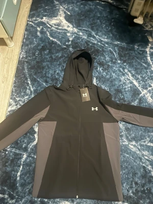 Svart vindjacka från Under Armour - Snygg svart vindjacka från Under Armour med gråa paneler på sidorna och ärmarna. Jackan har dragkedja framtill, justerbar huva och logga på bröstet. Perfekt för blåsiga dagar och sportiga outfits. Materialet är lätt och vindtätt.