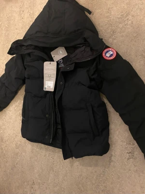 Svart dunjacka från Canada Goose (Macmillan) - Snygg svart dunjacka från Canada Goose med huva och klassisk logotyp på ärmen. Jackan har dragkedja och knappar framtill samt två stora fickor. Perfekt för kalla vinterdagar och riktigt varm tack vare dunfyllningen.