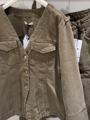 Gina figursydd jeansjacka - Snygg olivgrön jeansjacka med figurnära passform och markerad midja. Jackan har V-ringning, bröstfickor med lock och knappar framtill. Tydliga sömmar ger en cool vintagekänsla. Perfekt för dig som vill ha en unik twist på klassisk jeansjacka.