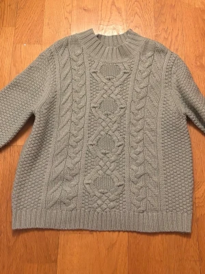 Grå kabelstickad tröja - Snygg grå stickad tröja med grovt kabelmönster framtill och ribbad hög hals. Tröjan har långa ärmar och breda muddar vid ärmslut och nederkant. Perfekt för kalla dagar och ger en klassisk look.