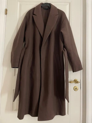 Brun lång kappa med knytskärp - Jättefin brun kappa i lång modell med knytskärp i midjan. Kappan har två fickor framtill och en rak, avslappnad passform. Använd endast någon enstaka gång och inte nopprig! Storlek S. OBS: Nypris är 800kr!!! 🤎