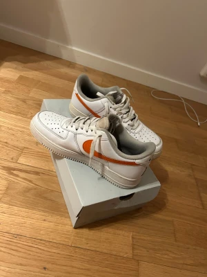 Nike Air Force 1 med orange swoosh - Fräscha Nike Air Force 1 sneakers i vitt läder med en unik swoosh i orange och gult på sidan. Klassisk siluett med perforerad tå, grå insida och vita snören. Perfekta för dig som vill sticka ut med en färgklick på fötterna. Ny pris på dessa skor när jag köpte de från amazon kosta de 4000kr