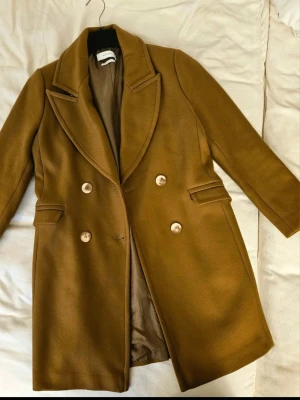 Kamelbrun dubbelknäppt rock - Snygg kamelbrun rock med dubbelknäppning och breda slag. Rocken har två fickor med lock och en klassisk rak passform. Tillverkad i ett ull-liknande material som ger en stilren och tidlös look. Perfekt för dig som vill ha en trendig och elegant ytterrock.