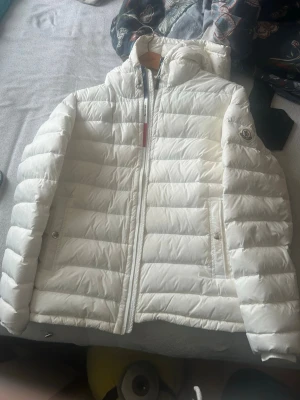 Vit dunjacka från Moncler med huva - Snygg vit dunjacka från Moncler med huva och dragkedja framtill. Jackan har två fickor med knappstängning och Moncler-logga på ärmen. Tillverkad i lätt, quiltat material som håller värmen under kalla vinterdagar.