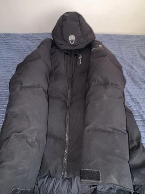 Svart Sail Racing pufferjacka med huva - Säljer en svart pufferjacka från Didriksons med stor vadderad huva och dragkedja framtill. Jackan har en bröstficka med dragkedja och kardborre vid ärmsluten. Perfekt för kalla vinterdagar och riktigt skön att bära.