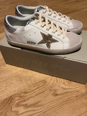 Golden Goose vita sneakers med glitterstjärna - Golden Goose sneakers i vitt läder med silverfärgade detaljer och en stor glitterstjärna på sidan. Grå mocka på tån och hälen, vita snören och låg profil. Snygg och unik design som sticker ut.