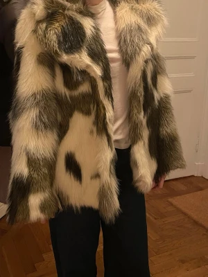 Fuskpälsjacka i beige och svart - Säljer en fluffig fuskpälsjacka med beige, svarta och bruna inslag. Jackan har bred krage och rak passform. Stängs framtill med tryckknappar. 