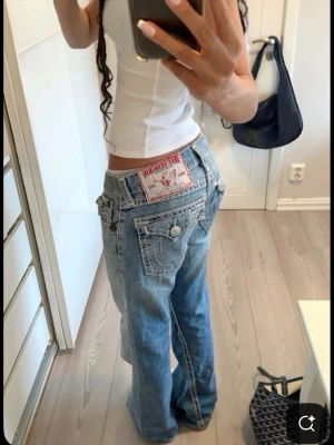 True Religion Joey jeans i ljusblå denim - Säljer ett par ikoniska True Religion Joey jeans i ljusblå denim med breda ben och låg midja. Jeansen har stora bakfickor med dekorativa sömmar och knappar, samt den klassiska True Religion-lappen bak. Perfekta för en avslappnad och trendig look.