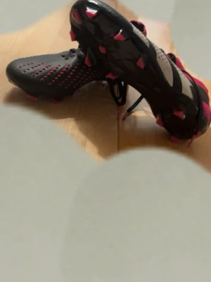 Adidas Predator Svarta Fotbollsskor - Snygga Adidas Predator fotbollsskor i svart med vita och röda detaljer. Skorna har snörning, låg profil och greppig sula med röda dobbar. Ovandelen är i syntetmaterial med prickigt mönster för bättre bollkontroll. Perfekta för dig som vill dominera på planen.