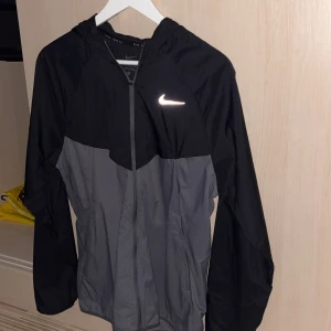 Nike svart och grå vindjacka - Snygg vindjacka från Nike i svart och grått med dragkedja framtill och huva. Kan byta imot en annan windbreaker helst Nike Miller svart eller Moncler eller annat d bara o visa.