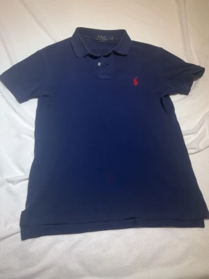 Marinblå pikétröja från Polo Ralph Lauren - Klassisk marinblå pikétröja från Polo Ralph Lauren med korta ärmar och krage. Tröjan har den ikoniska röda logotypen broderad på bröstet och knäppning vid halsen. Perfekt till jeans eller shorts för en clean och stilren look.