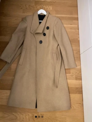 Beige ullkappa med knytskärp - Stilren beige ullkappa med dubbelknäppning och stora svarta knappar. Kappan har ett brett slag, två fickor framtill och ett matchande knytskärp i midjan. Lång modell som passar perfekt till kyliga dagar och ger en klassisk look.