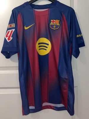 FC Barcelona matchtröja Lamine Yamal - Snygg FC Barcelona matchtröja i blått och rött med gula detaljer. Tröjan har klubbmärke, Nike-logga och Spotify som sponsor framtill. På ryggen står Lamine Yamal och nummer 10 i gult. Tillverkad i lätt och ventilerande material, perfekt för fotboll.
