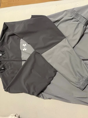 Grå sporttröja från Under Armour - Snygg sporttröja från Under Armour i två nyanser av grått. Tröjan har dragkedja framtill, lång ärm och logga på bröstet. Tillverkad i ett stretchigt och funktionellt material som passar perfekt för träning. Sitter som M 