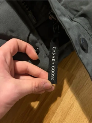 Mörkgrå dunjacka från Canada Goose - Snygg mörkgrå dunjacka från Canada Goose med klassisk svart logotyp på ärmen. Jackan har en stilren design och är tillverkad i slitstarkt material med dunfyllning som håller dig varm under kalla dagar.