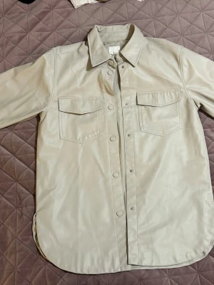 Beige overshirt i skinnimitation H&M - Stilren beige overshirt från H&M i skinnimitation. Modellen har två bröstfickor med lock, tryckknappar framtill och klassisk krage. Perfekt för lager-på-lager och ger en cool vibe till din outfit.