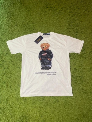 Vit Polo Ralph Lauren t-shirt med björn - Vit t-shirt från Polo Ralph Lauren med ikoniskt björntryck på framsidan. Klassisk passform och rund halsringning. Tillverkad i mjuk bomull och perfekt för en avslappnad stil.