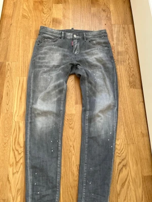 Grå Dsquared2 jeans med stänk - Säljer ett par grå jeans från Dsquared2 med coolt stänk- och slitningseffekt. Jeansen har smal passform, klassisk femficksdesign och en röd logotypdetalj fram. Materialet är mjukt denim i bomull. Bak finns en patch med texten 'Caten Heated' för extra attityd. knappt använda nästan helt nya inga skador eller något annat 