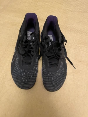 Svarta Reebok Nano sneakers - Snygga svarta Reebok Nano sneakers med lila detaljer och vit sula. Ovandelen är i mesh och syntet för bra andningsförmåga och komfort. Skorna har klassisk snörning och låg profil, perfekta för en sportig och modern look.