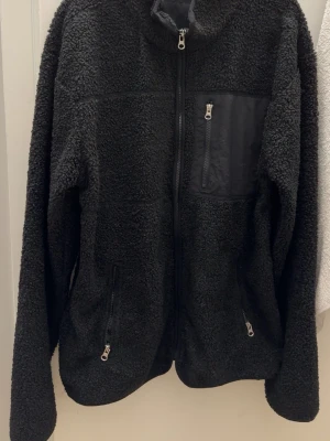 Svart teddyjacka med dragkedjor - Mysig svart teddyjacka med hel dragkedja framtill och tre fickor med dragkedja, varav en på bröstet. Jackan har hög krage och är tillverkad i fluffigt teddy-material som håller dig varm. Perfekt för kyliga dagar och enkel att matcha med olika outfits. Passar bra för M/L size