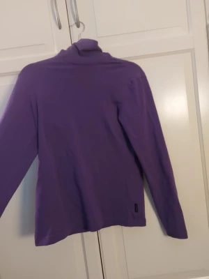 Lila tröja från Peak Performance - Säljer en lila långärmad tröja från Peak Performance i storlek M. Tröjan har hög krage och är tillverkad i ett mjukt och stretchigt material, perfekt för lager-på-lager. Enkel och stilren design utan tryck eller mönster.