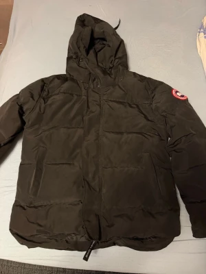 Canada Goose jacka  - Säljer en svart dunjacka från Canada Goose med huva och klassisk logotyp på ärmen. Jackan har två sidofickor med dragkedja och en hög krage. Perfekt för kalla vinterdagar och riktigt varm tack vare dunfyllningen. Hör av er för mer info.