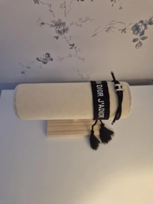 Elegant Dior armband set (2 st) - Snyggt svart tygarmband från Dior med texten 'J'ADIOR' broderad i vitt. Armbandet har justerbar knytning och dekorativa tofsar i ändarna. Perfekt accessoar för att lyfta din outfit med en exklusiv touch.