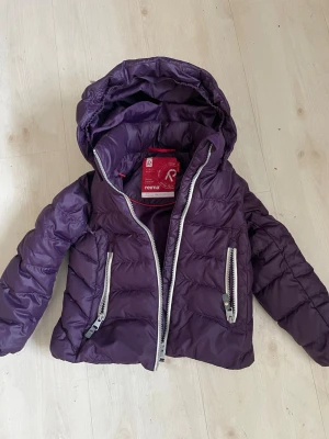 Lila pufferjacka från Reima - Snygg lila pufferjacka från Reima med avtagbar huva och två stora dragkedjefickor framtill. Jackan är vadderad och har en glansig finish, perfekt för kalla vinterdagar. Materialet är slitstarkt och håller värmen bra.