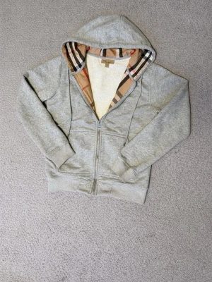 Grå hoodie med Burberry-mönster - Säljer en grå zip-hoodie från Burberry med klassiskt rutigt foder i huvan och insidan. Hoodien har dragkedja framtill, ribbade muddar och fickor på sidorna. Materialet är mjuk bomullsblandning och den har en avslappnad passform. Perfekt för dig som gillar stilrena detaljer.