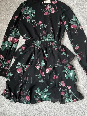 Svart blommig klänning från Ü - Säljer en svart klänning med blommigt mönster från Ü. Klänningen har långa ärmar och en volangdetalj vid kjolen.inga tecken på användning🩷